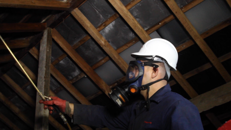 A technician in a boiler suit, hardhat and gas mask applying woodworm spray to timbers in loft